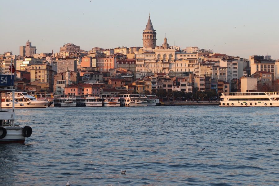 View across calm water of Istanbul cityscape, with Galata Tower standing above the other buildings in the center of the frame. It's the golden light hour as sun strikes the buildings.