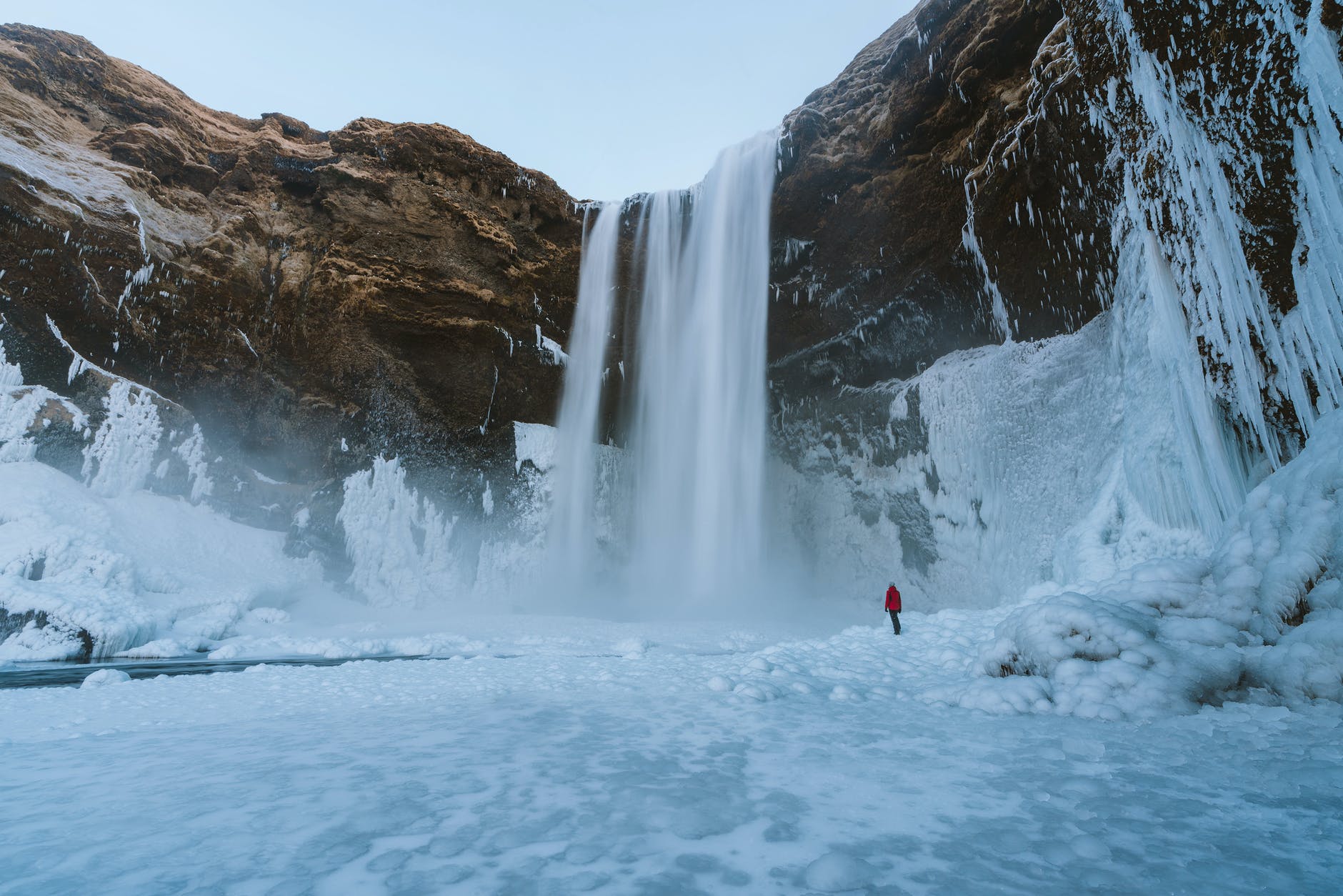 A person in a red coat in the distance in front of a tall waterfall amidst a snowy, white landscape. | Plan a trip to Iceland with these tips from Wanderful