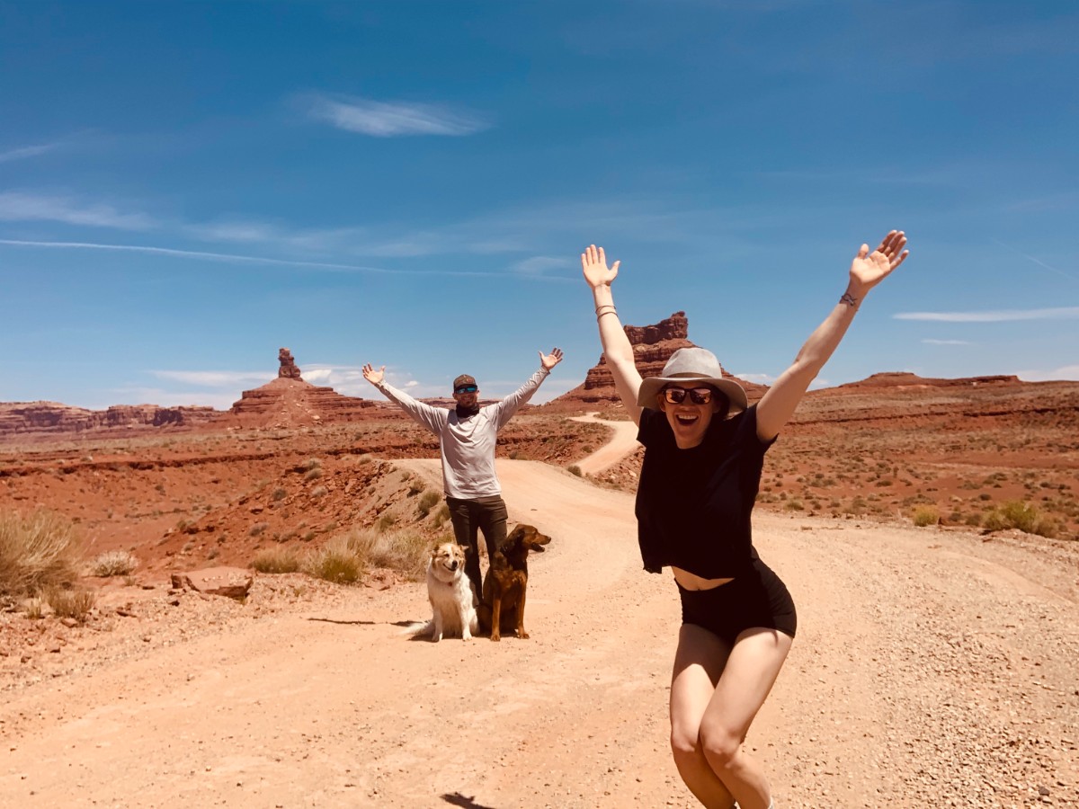 Two joyful people posing with arms outstretched overhead, with two dogs, all seemingly in the middle of a large desert