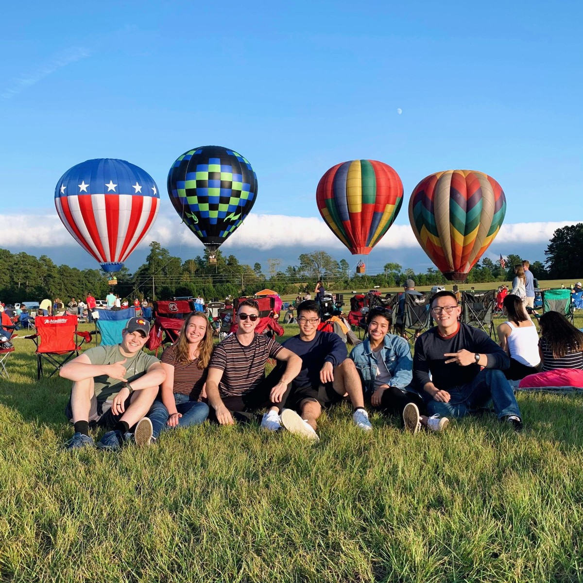 Group of friends sitting on a grassy hill with hot air balloons in the background