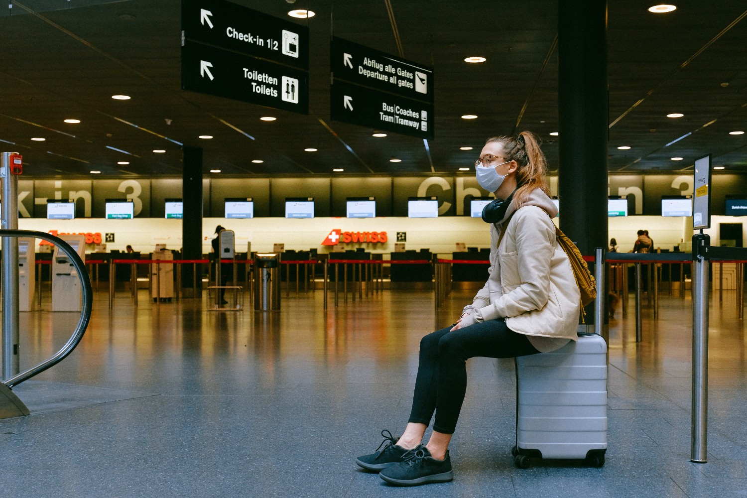 Woman sitting on a rolling suitcase in an airport wearing a face mask to travel in this new world