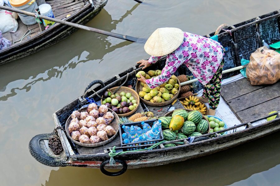A lady on a small boat in water. There are baskets of fruit such as watermelons, pineapples, and small bananas in the boat too.