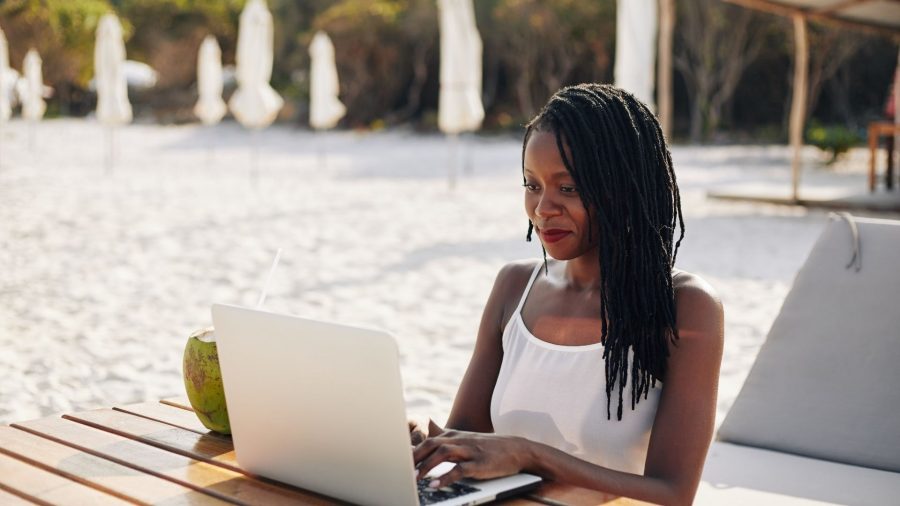A woman on the beach, using a laptop drinking coconut from a coconut.