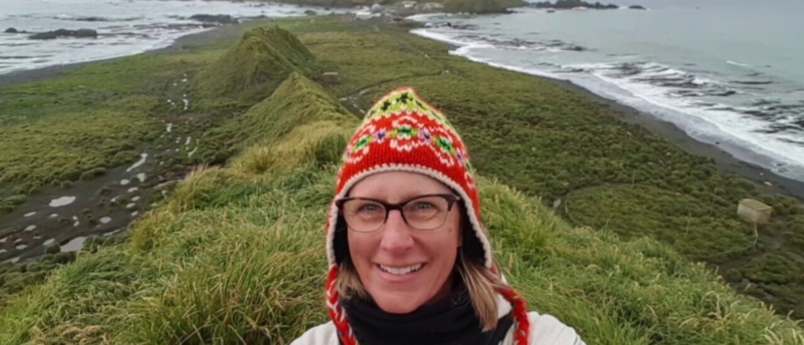 Sherry Ott wearing a colorful knitted hat and taking a selfie with a background of a green strip of land with the ocean on both sides - presentation for Wanderful titled Where to Go & When