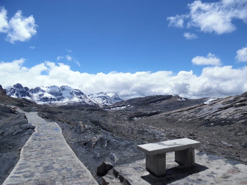 A mountaintop vista with blue sky and clouds on the horizon, and a small bench in the foreground