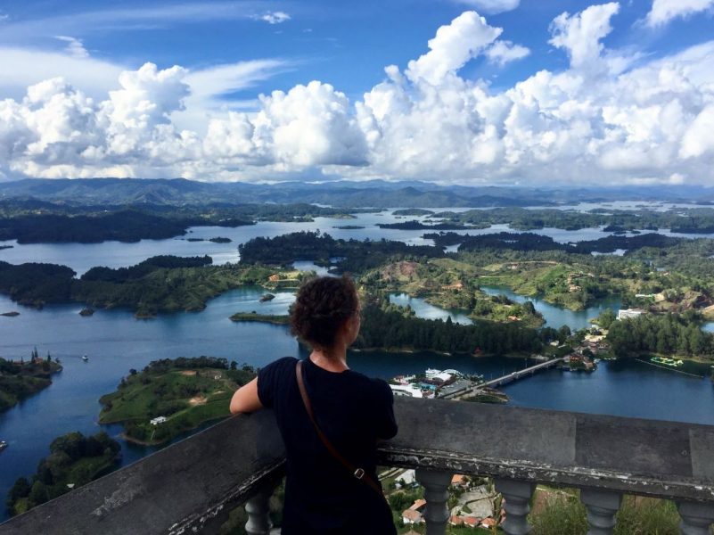 A woman overlooks the Piedra de Penol in Colombia from a patio vantage point high above various lakes and mountains on the horizon