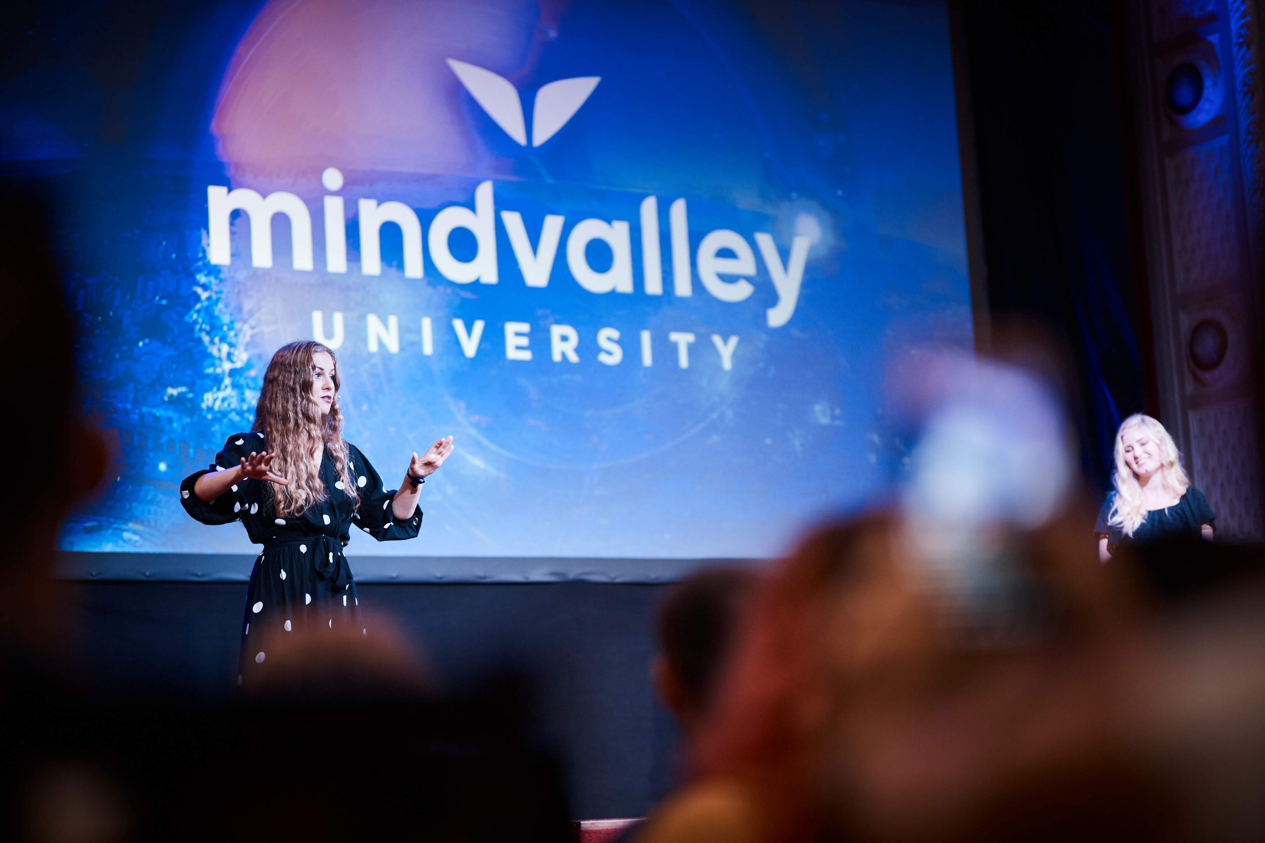Kaitlin Murray of Odyssey HQ speaking on stage with the MindValley University logo behind her | Wanderful