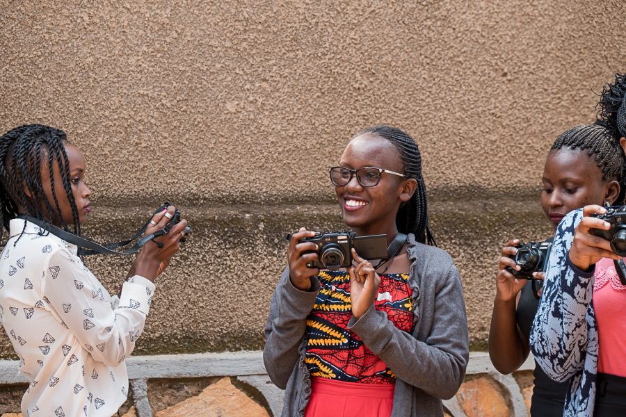 A young woman holds a camera smiling as several women nearby practice on their own cameras, part of the Cameras for Girls program in Uganda