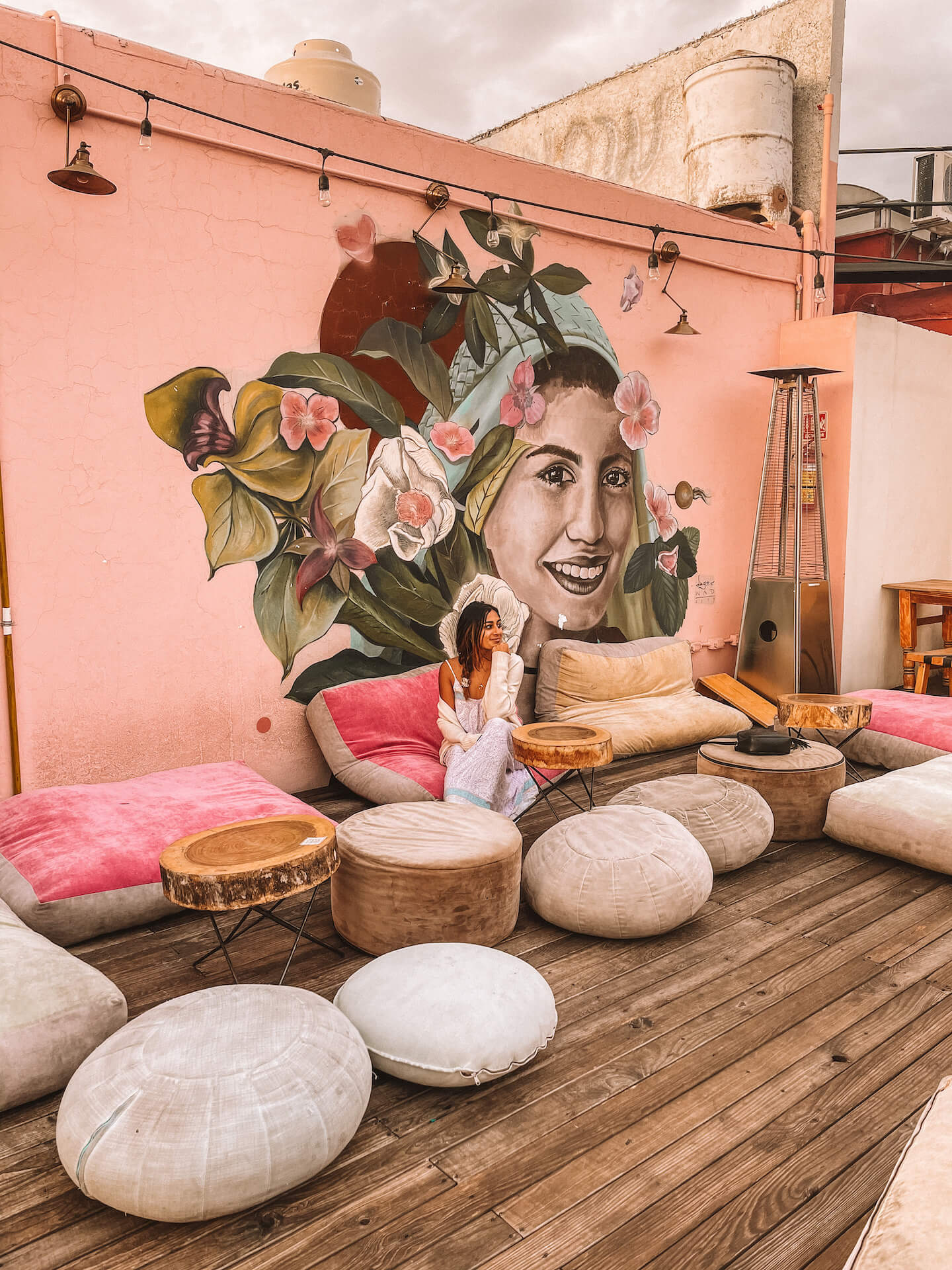 Nabila Ismail sits amidst a plush outdoor patio with a bright mural on the pink wall behind her