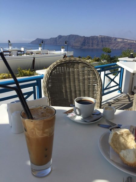 A table with coffee and tea on it. Wicker chair and a view of a boat, the sea and cliffs.