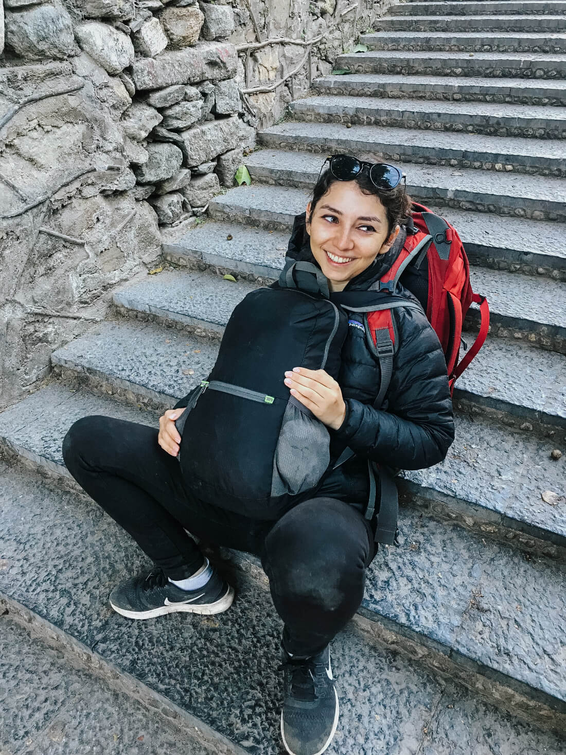 Stacey Valle with a large backpack and another smaller backpack on her front, sitting on a staircase looking exhausted but smiling